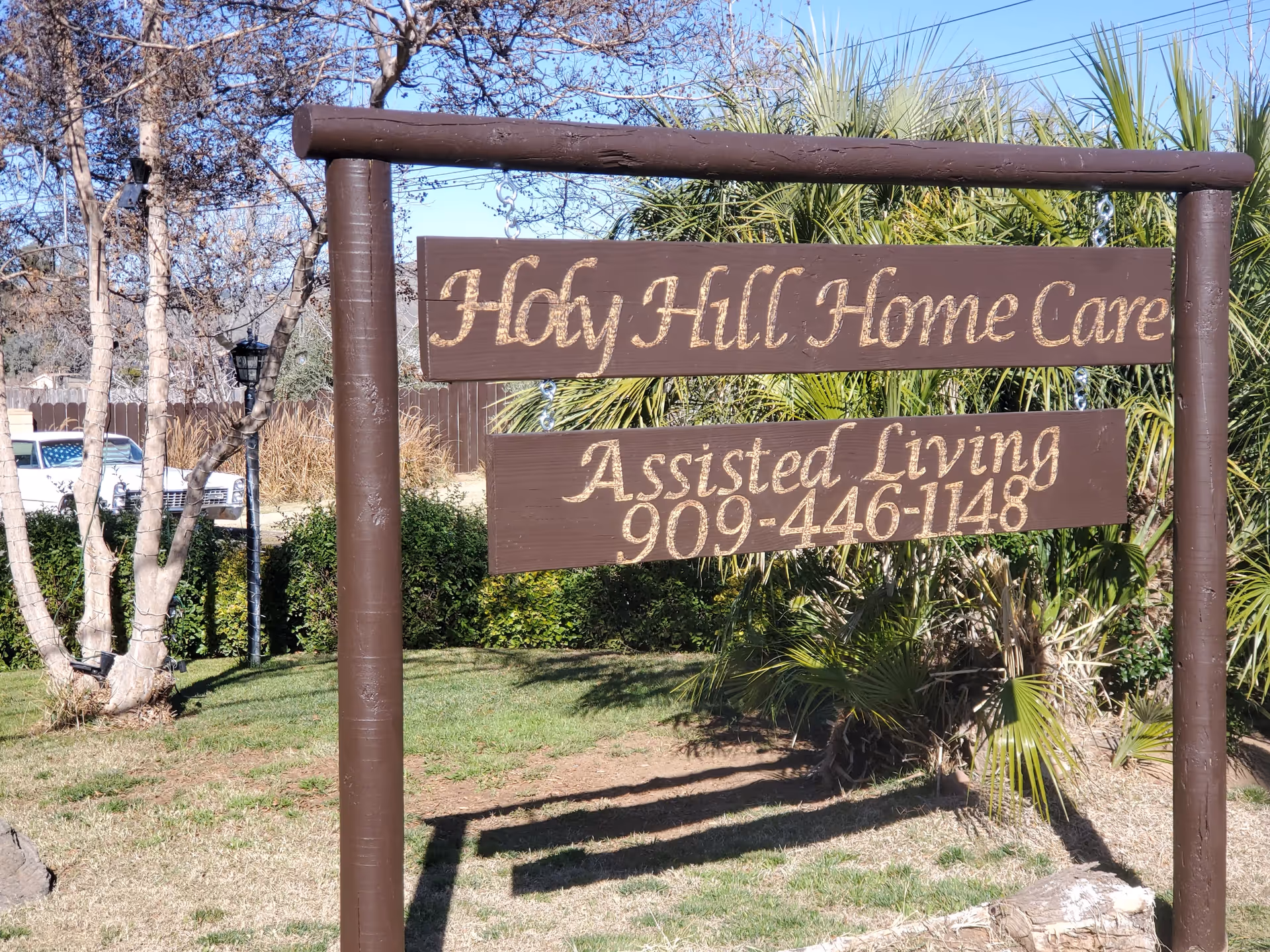 Wooden roadside sign reading "Holy Hill Home Care" and "Assisted Living 909-446-1148" set in a grassy yard with trees and shrubs behind it.