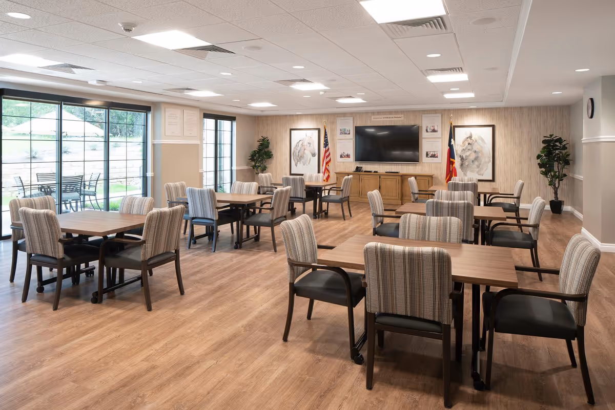 A spacious senior living facility common room with multiple wooden tables and cushioned chairs arranged neatly. Large windows and glass doors on the left side provide natural light and a view of an outdoor patio with tables and chairs. The room features wood flooring, a large flat-screen TV mounted on the far wall, two framed horse paintings, and two flags on either side of the TV. Potted plants are placed in the corners, and the ceiling has recessed lighting.