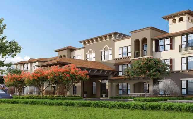 Exterior view of a multi-story senior living facility building with a covered entrance, surrounded by green grass and trees with red flowers under a clear blue sky.