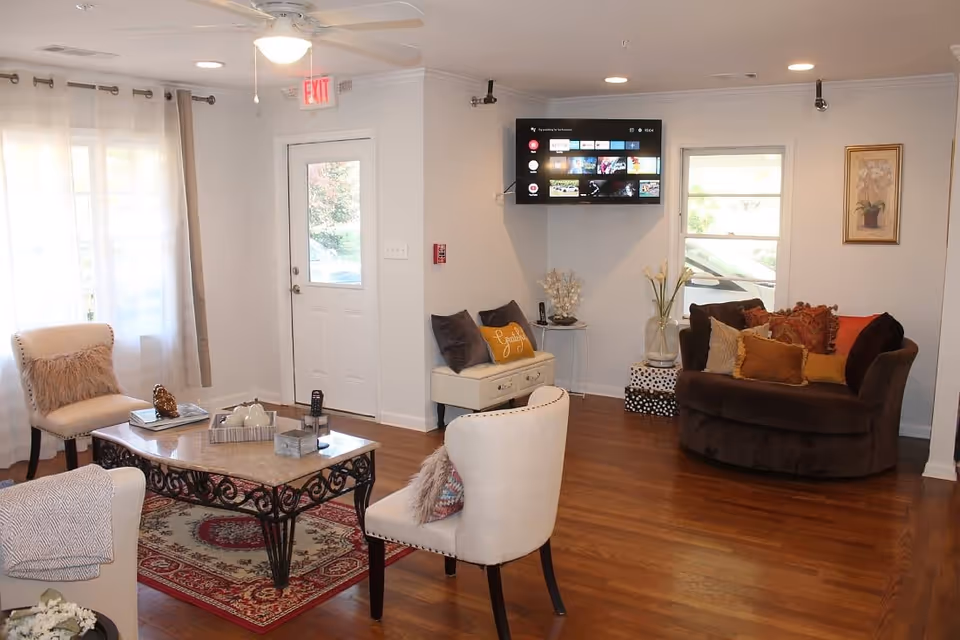 A cozy living room with wooden floors, a ceiling fan, and a wall-mounted flat-screen TV. The room features a brown cushioned loveseat with multiple pillows, two beige chairs with decorative pillows, a coffee table with decorative items, and a small bench with pillows. There are windows with curtains and a door with a window, allowing natural light to enter the space.