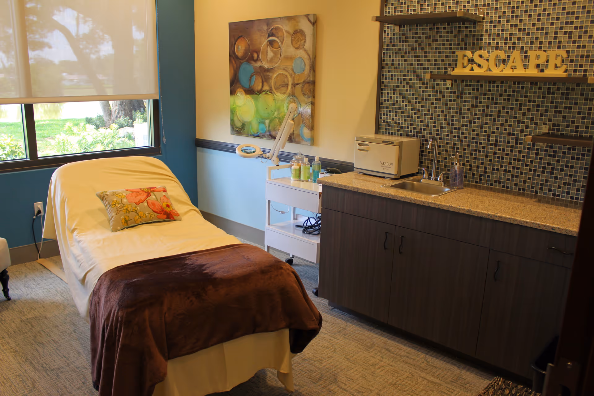 A cozy room with a reclining chair covered with a white sheet and a brown blanket, a floral pillow on the chair, a window with a view of greenery outside, a countertop with a sink and cabinets, a small cart with bottles and equipment, a colorful abstract painting on the wall, and the word ESCAPE displayed on a mosaic tile backsplash.