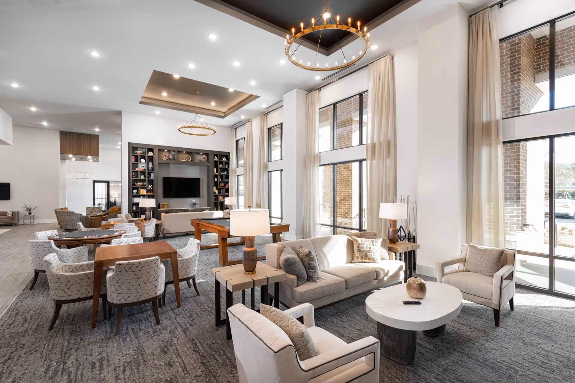 A spacious and well-lit living room area in a senior living facility with large windows covered by sheer curtains. The room features multiple seating arrangements including beige sofas and armchairs around a white oval coffee table, wooden tables with patterned chairs, and a pool table. There are two large circular chandeliers hanging from the ceiling and a built-in entertainment center with a flat-screen TV and decorative shelves.