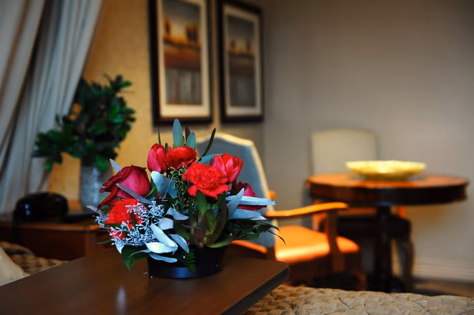 A close-up of a vibrant floral arrangement with red flowers and green leaves placed on a wooden table in a cozy living room setting. In the background, there are two framed pictures on the wall, a wooden round table with a bowl on it, and two chairs with armrests.