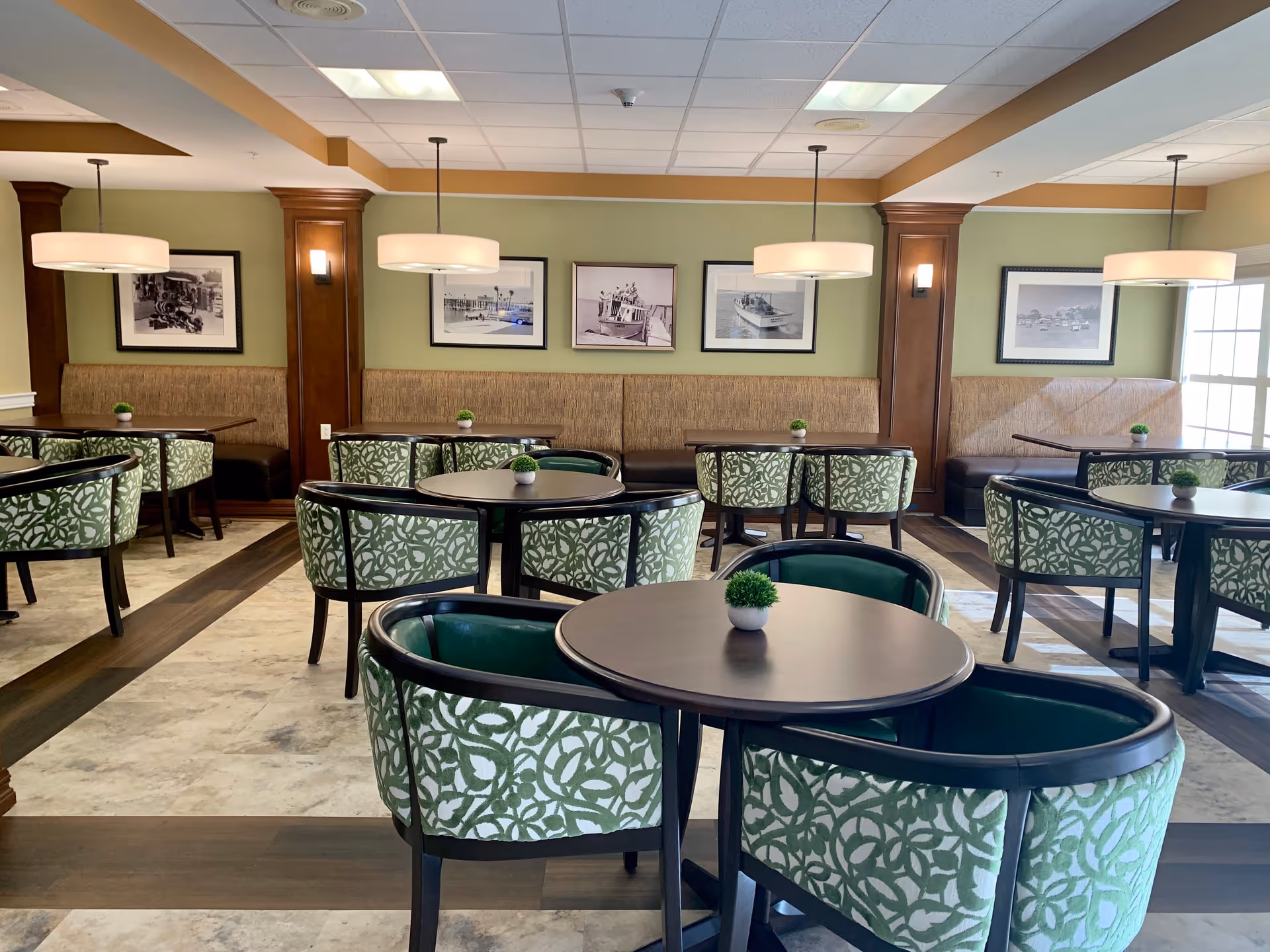 A dining room in a senior living facility with round dark wood tables surrounded by green patterned chairs. The walls are painted light green with wooden columns and framed black and white photographs. Pendant lights hang from the ceiling, and small potted plants are placed on each table.