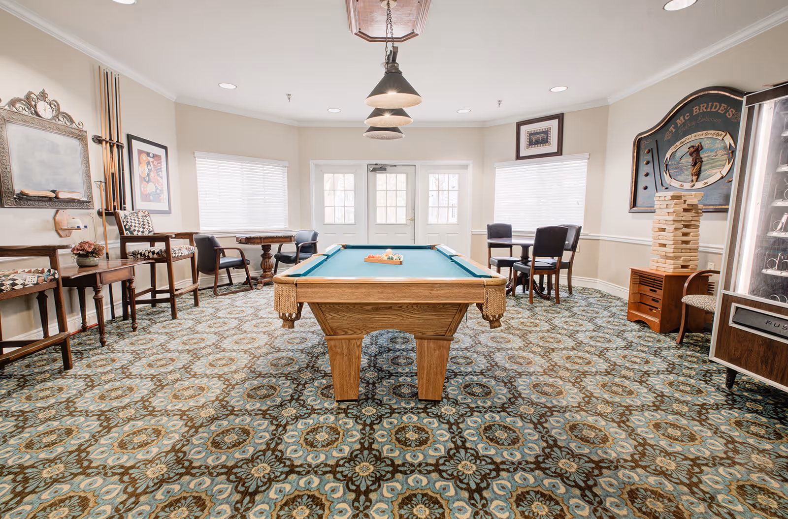 A recreational room with a pool table in the center, surrounded by chairs and small tables. The room has patterned carpet, beige walls, and several windows with blinds. There are hanging lights above the pool table, a large Jenga game on a small cabinet, and framed artwork on the walls.
