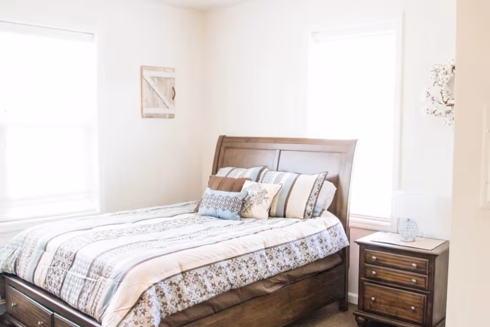 Bright bedroom with a wooden bed, patterned bedding, and a nightstand between two windows.