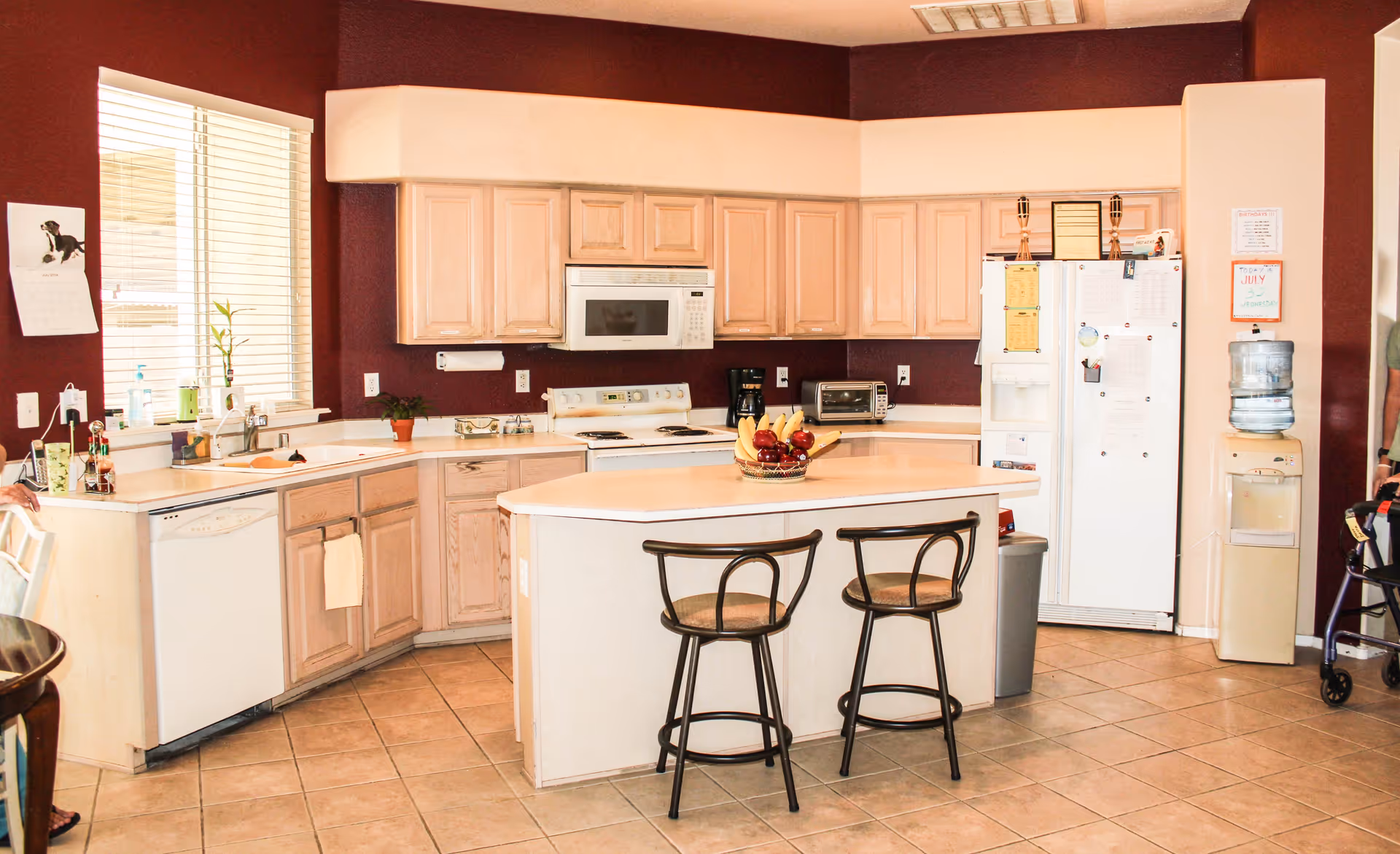 A kitchen with light wood cabinets and a white countertop island with two bar stools. The kitchen features a white stove with a microwave above it, a dishwasher, a refrigerator covered with notes and magnets, a water cooler, and a tiled floor. There is a window with blinds on the left wall and a basket of fruit on the island.