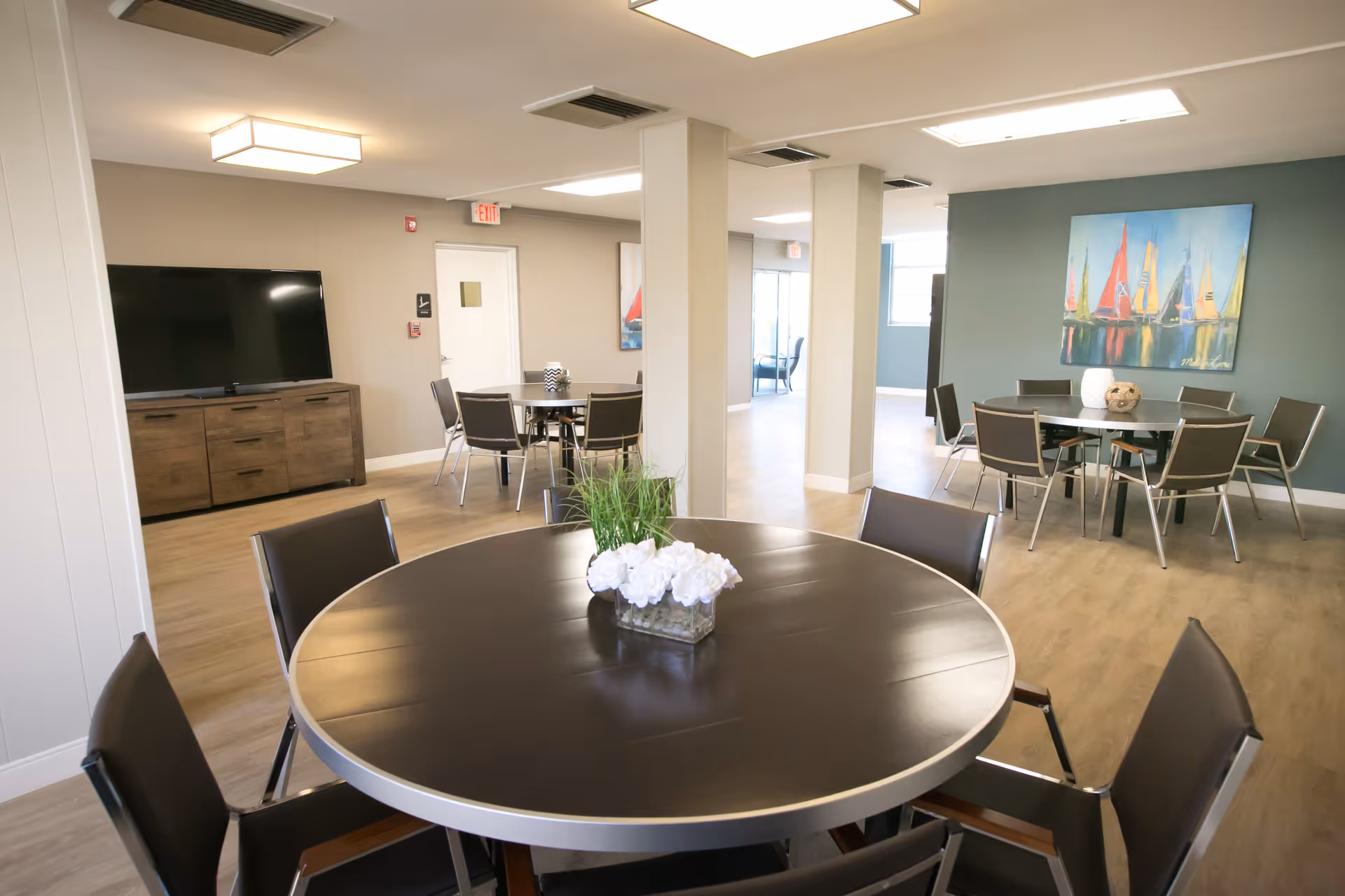 A bright and spacious common area with multiple round tables surrounded by chairs. Each table has a decorative centerpiece with white flowers and greenery. The room features light wood flooring, beige and teal walls, a large flat-screen TV on a wooden cabinet, and colorful sailboat artwork on the wall. Ceiling lights illuminate the space, and there are visible exit signs and doors in the background.