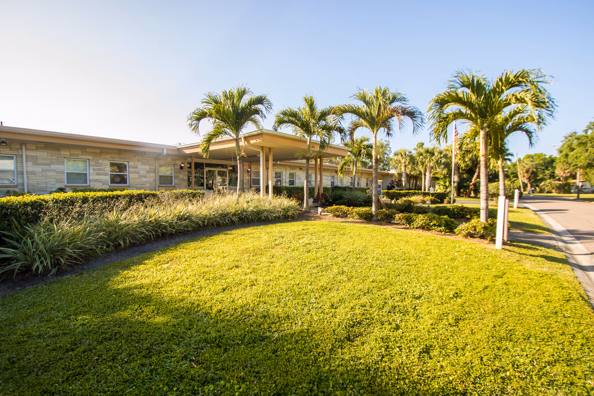 Exterior view of Shore Acres Care Center building with a well-maintained lawn, palm trees, and shrubs under a clear blue sky.