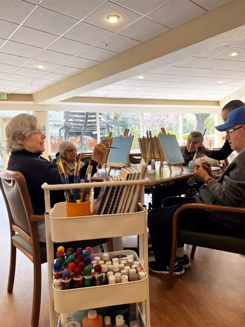 A group of elderly individuals seated around a table in a well-lit room, engaged in painting on canvases placed on easels. In the foreground, there is a cart filled with paintbrushes, paint bottles, and art supplies. Large windows in the background let in natural light and show some outdoor greenery.