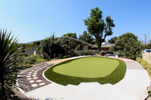 Outdoor area at Upland Rehabilitation & Care Center featuring a putting green surrounded by landscaped plants, trees, and a paved walking path under a clear blue sky.