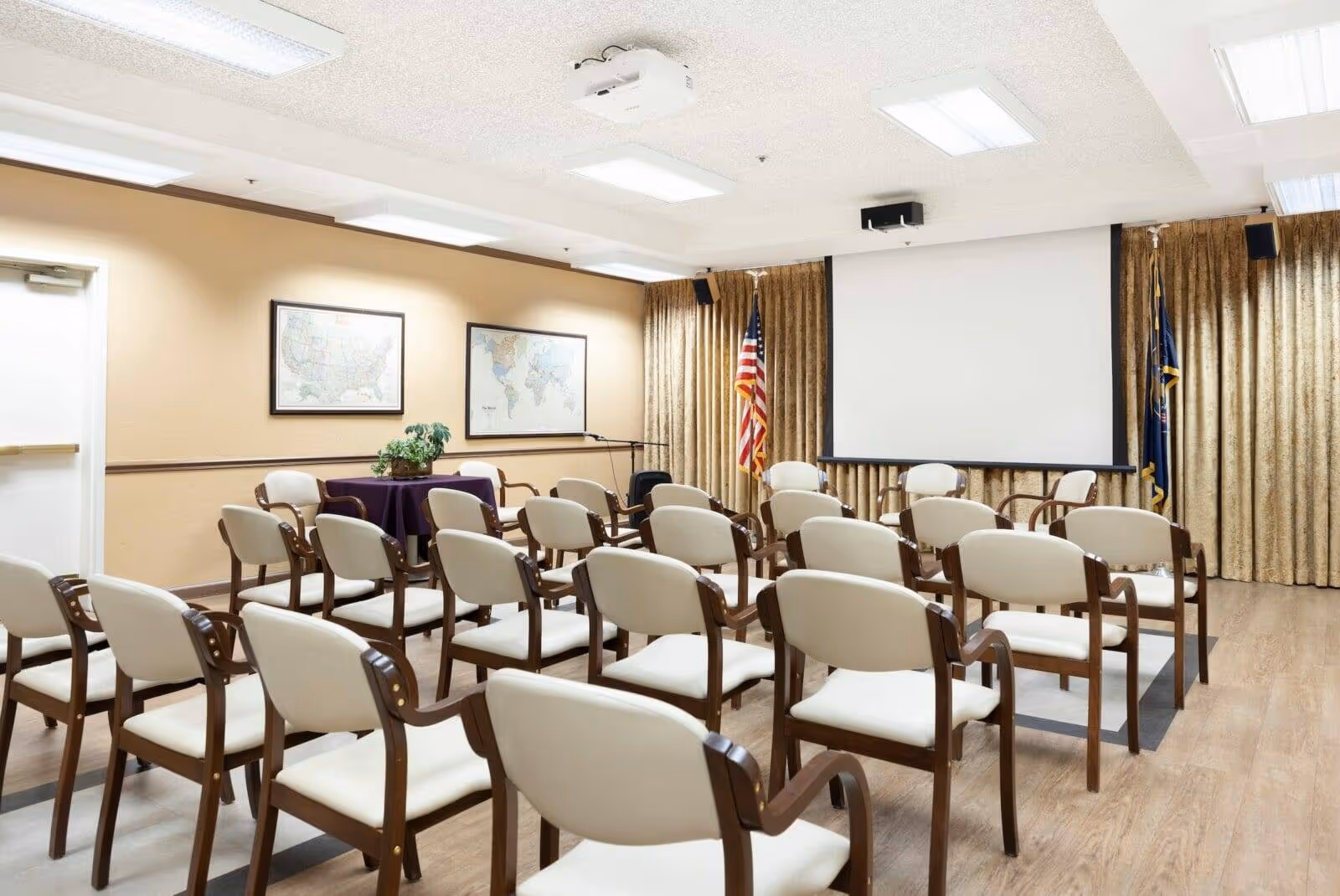 A conference or meeting room with rows of beige cushioned chairs with wooden armrests facing a large white projection screen. The room has beige walls with two framed maps, a table with a purple tablecloth and a plant, and American and state flags near the screen. The ceiling has fluorescent lights and a projector mounted.