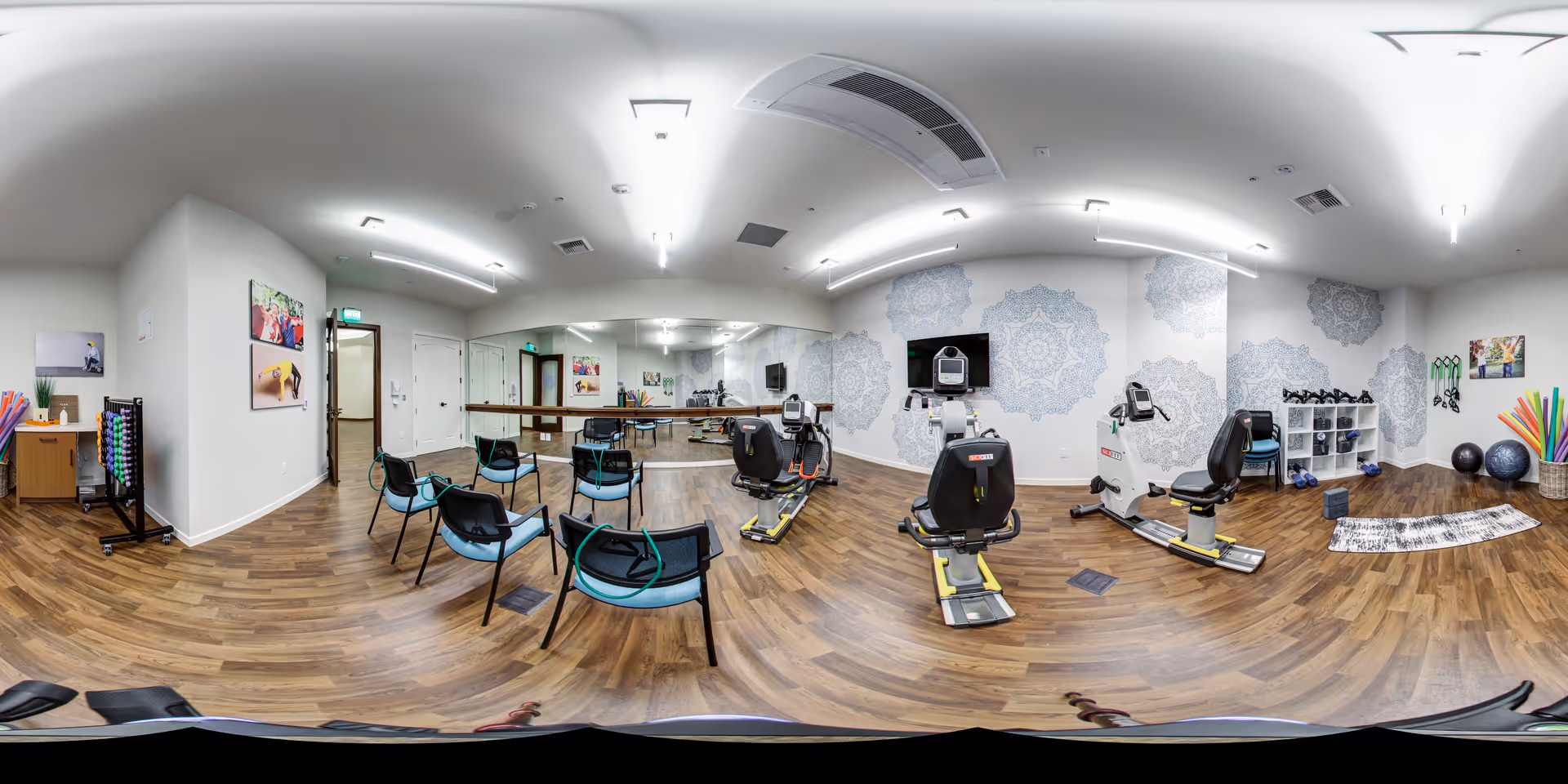 Bright fitness/activity room with recumbent exercise bikes, rows of chairs, mirrors, and shelved workout equipment on wood floors.