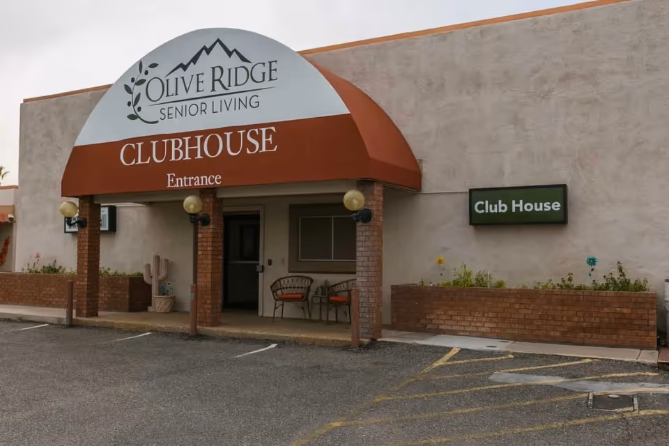 Exterior view of the Olive Ridge Senior Living clubhouse entrance with a red awning and brick pillars, two chairs and a small table near the entrance, and a parking area in front.