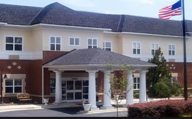 Exterior view of The Living Center of Concord, showing a two-story building with a covered entrance supported by white columns, an American flag on a flagpole, and landscaping with bushes and small trees.