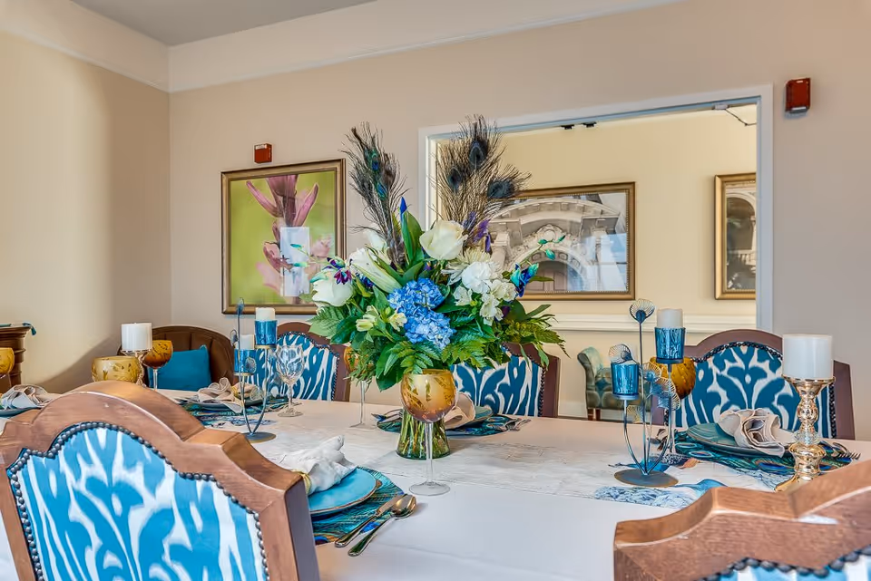 A dining table set with blue and gold glassware, beige napkins, and a centerpiece floral arrangement featuring white roses, blue hydrangeas, and peacock feathers. The chairs around the table have wooden frames with blue patterned upholstery. The room has beige walls with framed artwork and a window opening to another room with additional seating.