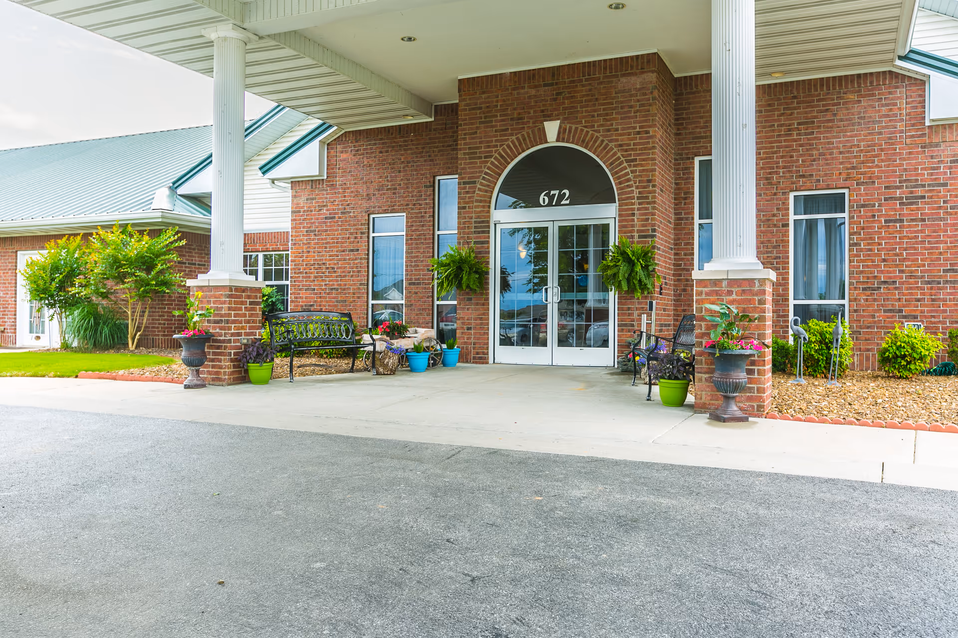 Entrance of a brick building with the number 672 above double glass doors. The entrance is covered by a roof supported by white columns. There are benches and various potted plants arranged around the entrance area.