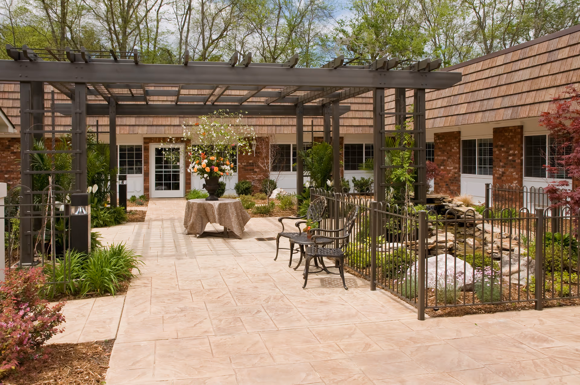 Outdoor courtyard area with a pergola, metal benches, a table with a floral arrangement, and landscaped garden beds surrounded by a low metal fence. The courtyard is adjacent to a brick building with multiple windows and doors.