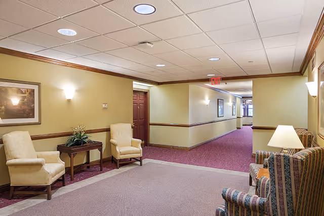 A well-lit hallway in a senior living facility with beige walls, maroon carpet, and ceiling lights. The hallway features two beige upholstered armchairs with a small wooden table and plant between them on the left side, and two striped upholstered armchairs with a lamp on a side table on the right side. There are framed pictures on the walls and an exit sign visible in the distance.