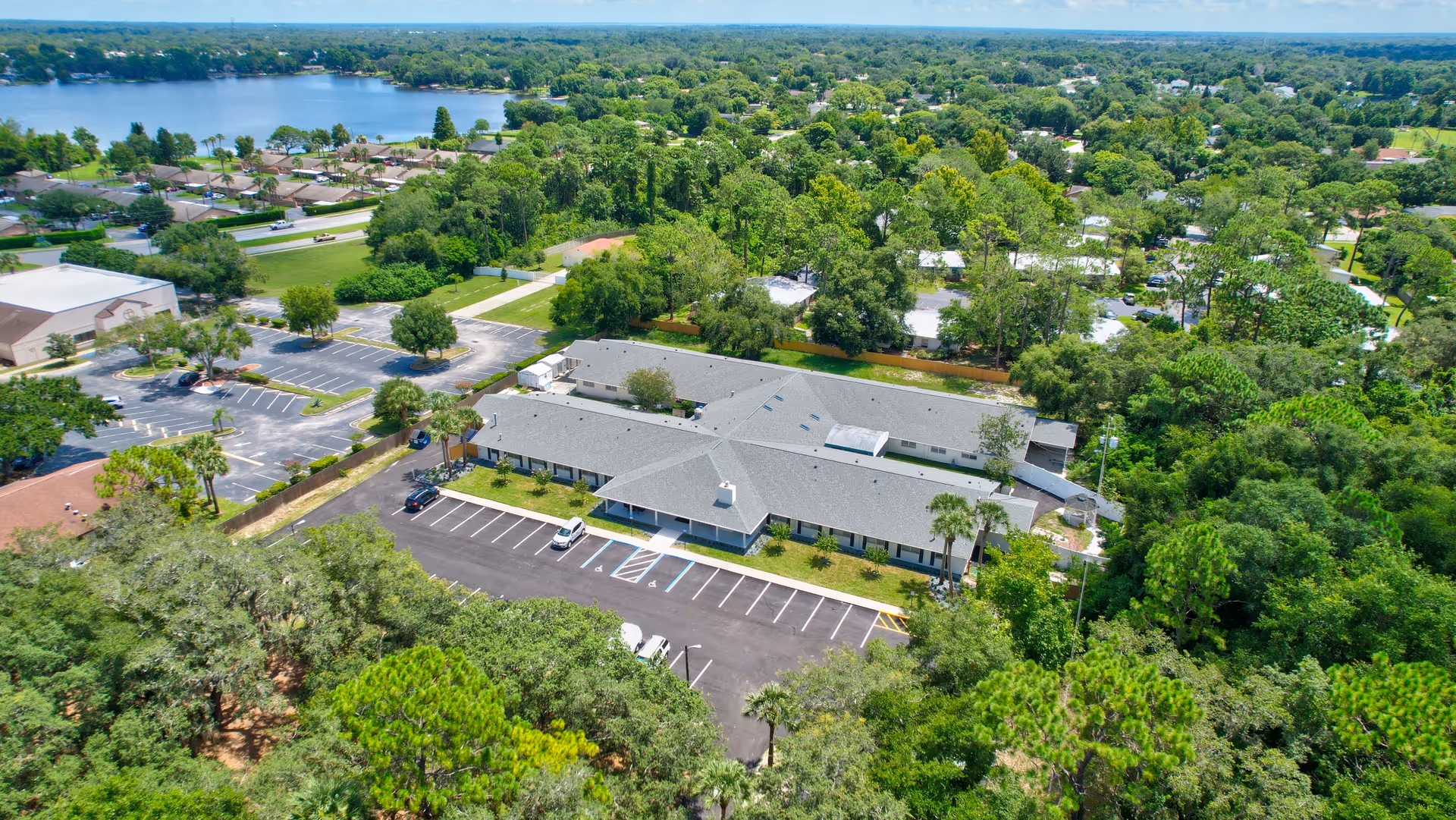 Aerial view of The Gardens of Eastbrooke facility surrounded by trees and greenery, with a parking lot in front and a lake visible in the background under a clear blue sky.