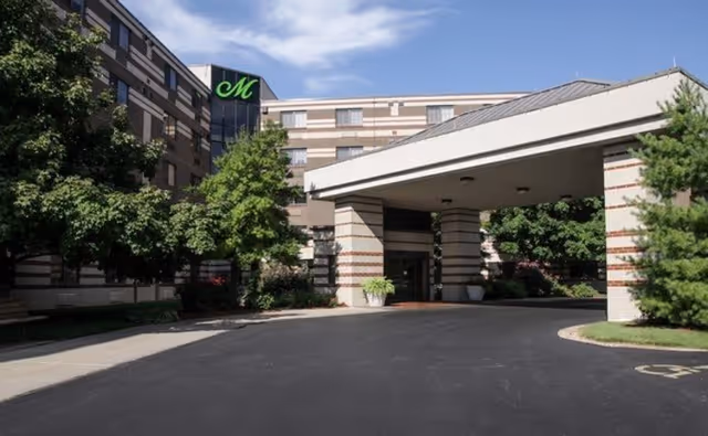 Exterior view of The Montclair Senior Living facility showing the main entrance with a covered driveway, surrounded by trees and landscaping under a partly cloudy sky.