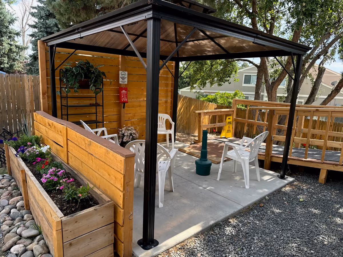 Outdoor seating area with a black metal gazebo structure and wooden privacy walls. Inside the gazebo are several white plastic chairs arranged around a green umbrella stand. There is a wooden planter box with colorful flowers along the left side, and a wooden ramp with railings leading to the area. Trees and a wooden fence surround the space.