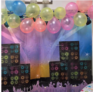 Colorful party decorations with clusters of pastel balloons in blue, green, pink, and yellow hanging above a backdrop featuring a vibrant, abstract design with silhouettes of cheering people and stacked speaker illustrations.