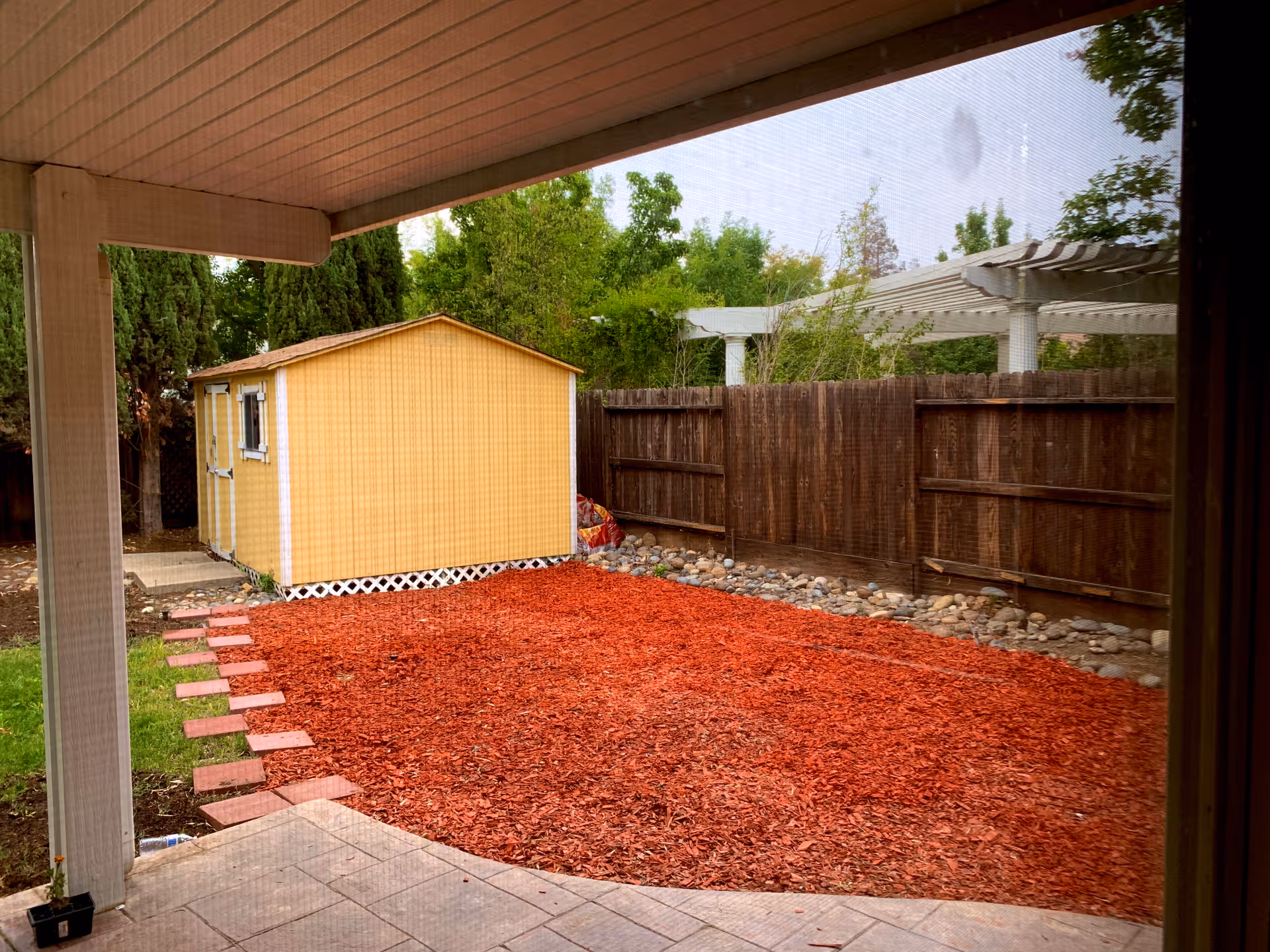 View of a backyard area with a covered patio in the foreground, a red mulch ground covering, a small yellow shed, a wooden fence, and green trees in the background.