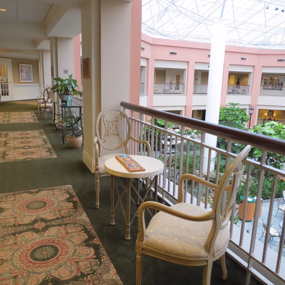 Indoor balcony area overlooking a large atrium with greenery and seating below. The balcony has a green carpet with patterned rugs, cream-colored chairs, a small round table with a board game on it, and potted plants along the railing and walls. The ceiling is a glass dome allowing natural light to fill the space.