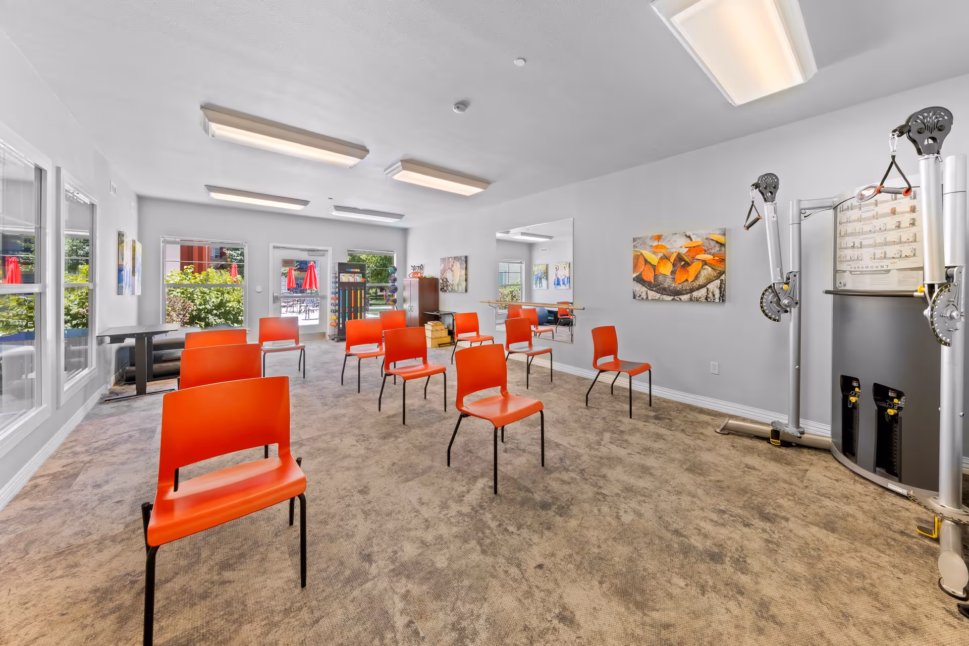 A bright room with several orange chairs arranged in rows on a carpeted floor, a large mirror on the wall, exercise equipment on the right side, and windows and a door leading outside with visible greenery and red umbrellas.
