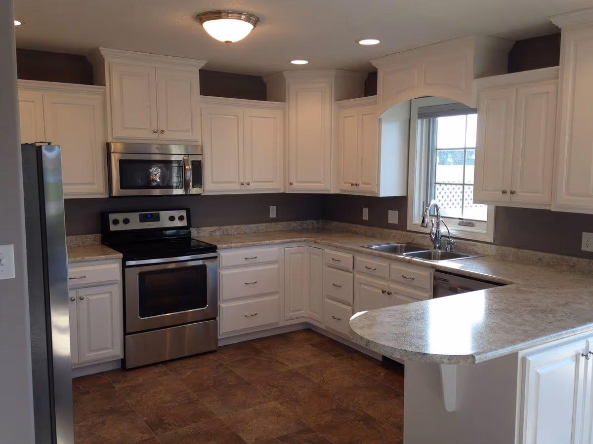 A modern kitchen with white cabinets, stainless steel appliances including a refrigerator, oven, and microwave. The kitchen has a double sink under a window, granite countertops, and a tiled floor.