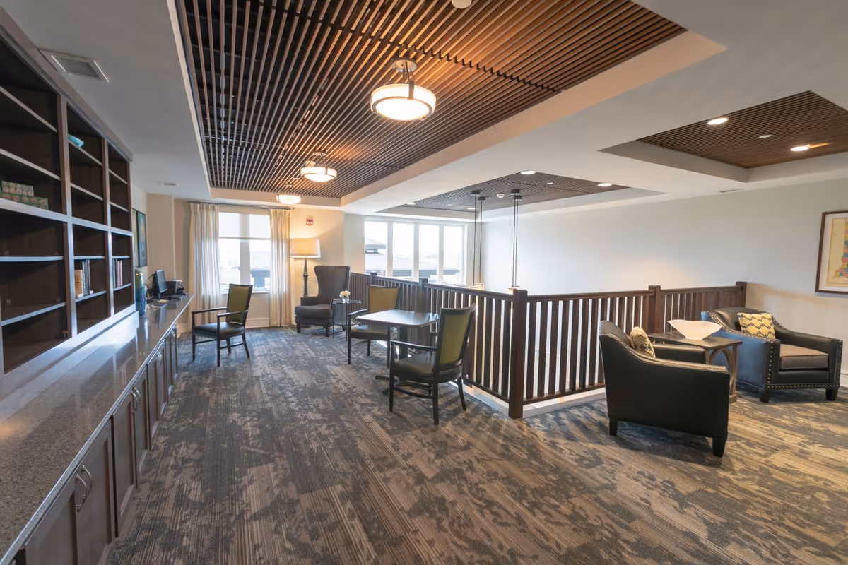 A spacious, well-lit common area with modern decor featuring a patterned carpet, wooden ceiling panels with hanging lights, and large windows with curtains. The room includes several seating arrangements with chairs and small tables, a tall floor lamp, and built-in wooden shelves along one wall.