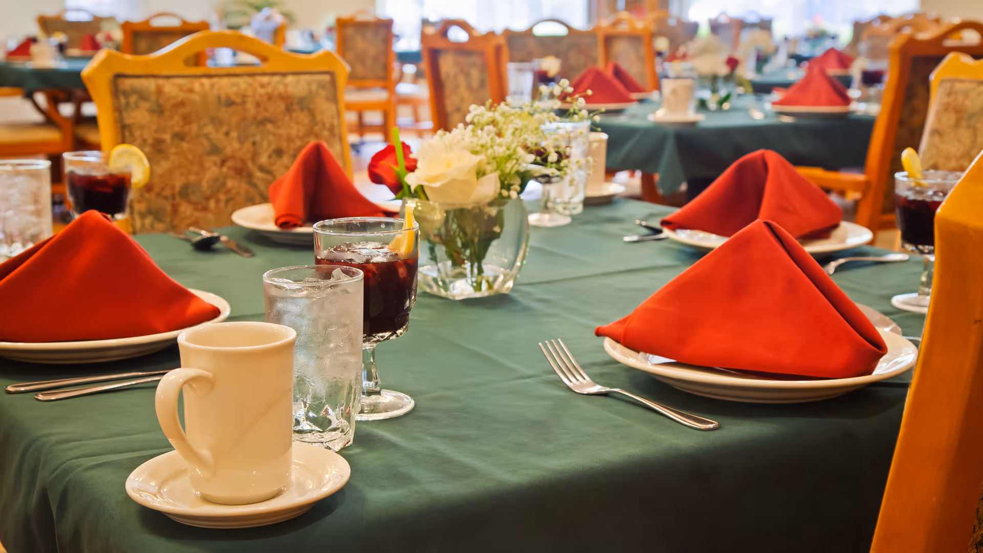 A dining table set with green tablecloth, white plates with red folded napkins, glasses of water and iced tea with lemon, silverware, and a small floral centerpiece in a clear vase. The background shows more similarly set tables and chairs in a dining room.