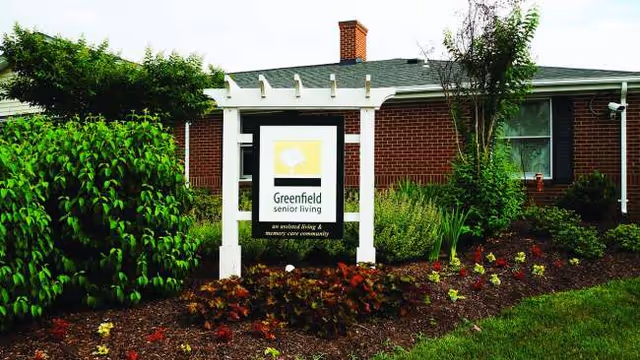 Exterior view of a brick senior living building with a white pergola and landscaped yard