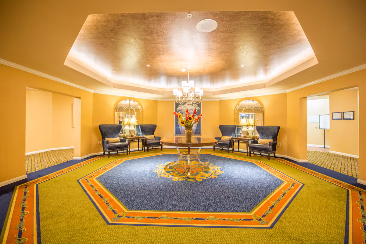 A spacious, well-lit interior room with a large octagonal carpet featuring blue, yellow, and orange patterns. In the center of the room is a round wooden table with a vase of colorful flowers. Surrounding the table are four dark blue upholstered armchairs with small side tables and lamps behind them. The walls are painted yellow, and there are two large arched mirrors on the back wall. The ceiling has a recessed design with warm lighting and a chandelier hanging above the table.