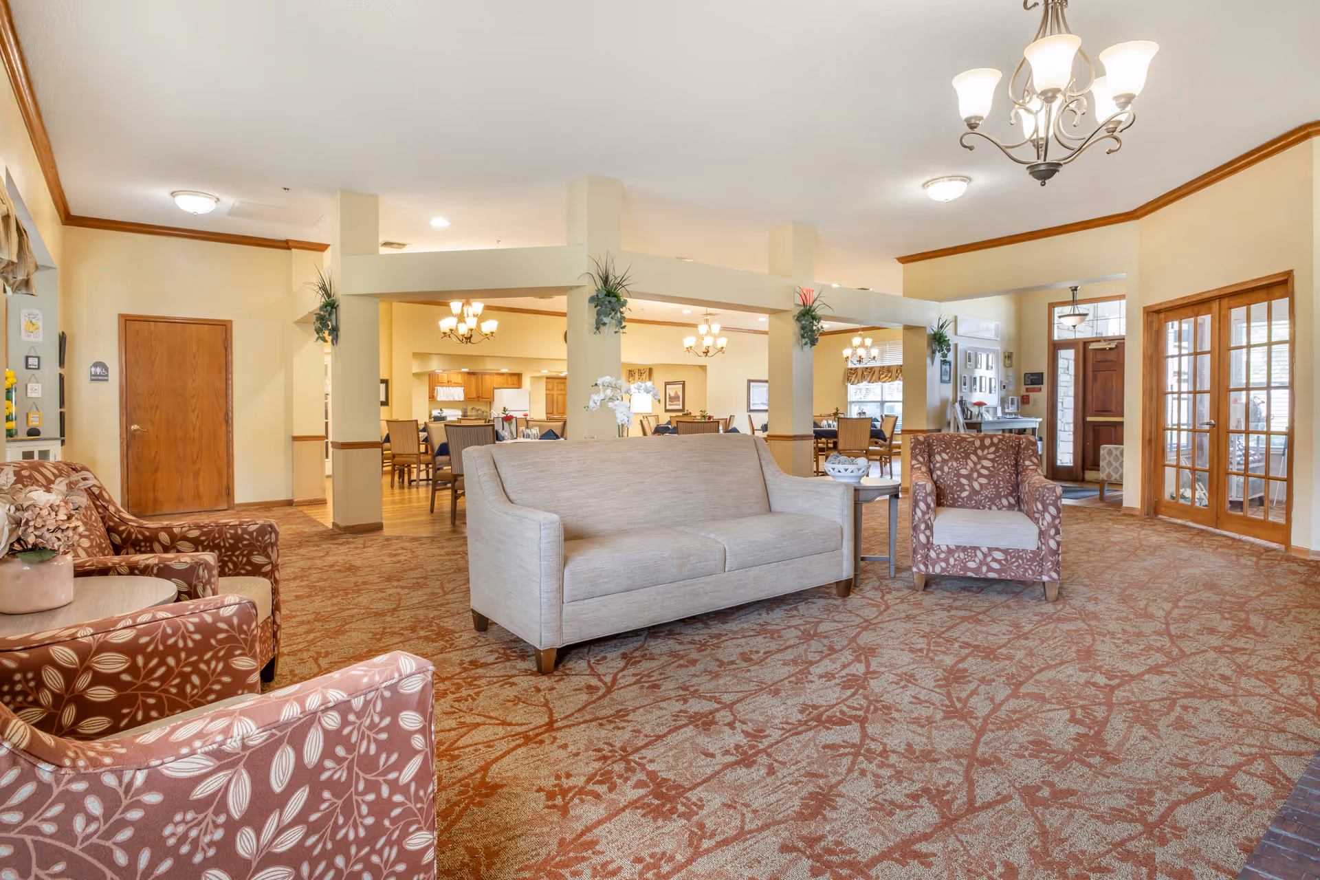 A spacious senior living facility common area with beige walls and patterned carpet. The room features a beige sofa and two floral-patterned armchairs arranged around a small table with a flower vase. In the background, there is a dining area with multiple tables and chairs, wooden cabinetry, and warm lighting fixtures. The entrance door with glass panels is visible on the right side.