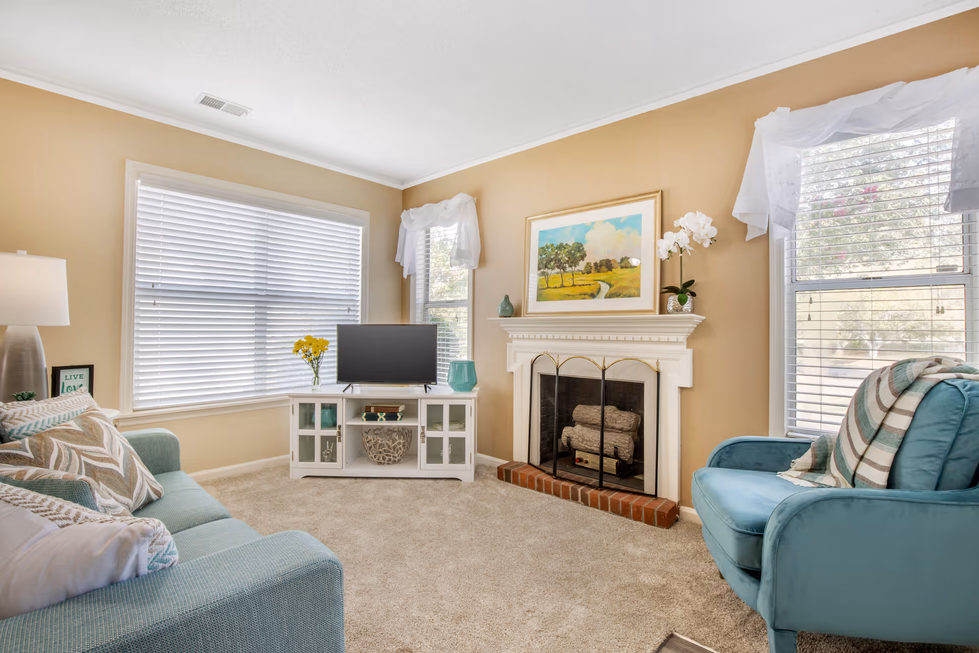 A cozy living room with beige walls and carpeted floor. The room features a teal sofa with patterned cushions on the left, a teal armchair with a striped throw blanket on the right, and a white TV stand with a flat-screen TV and decorative items in the center. There is a white fireplace with a brass screen and a landscape painting above it. Two windows with white blinds and sheer white curtains let in natural light.