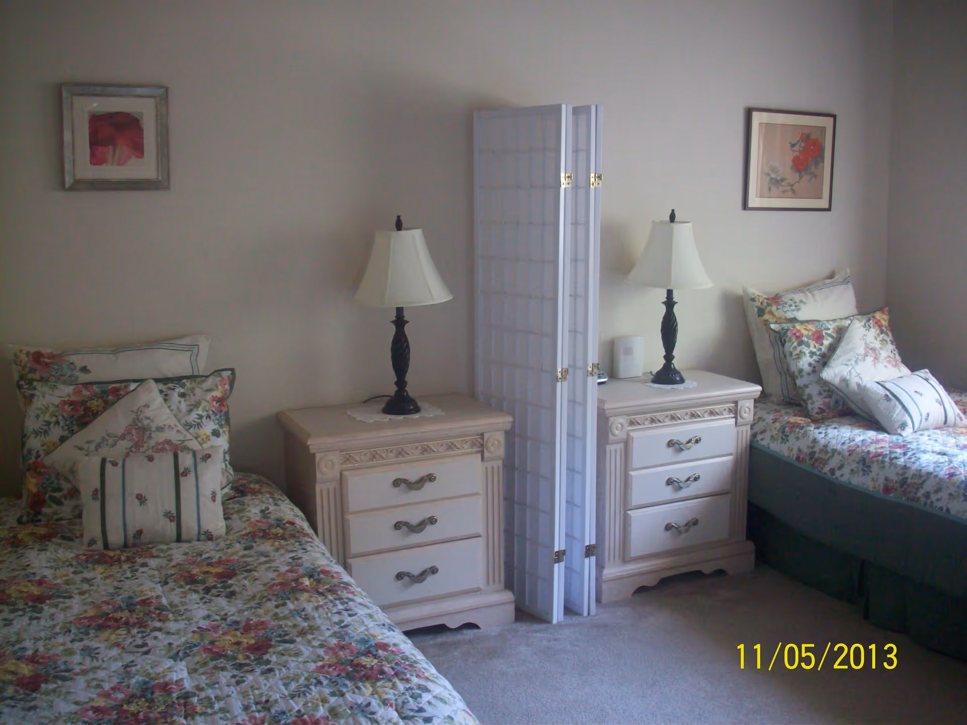 A bedroom with two twin beds separated by a white folding screen. Each bed has floral-patterned bedding and multiple pillows. Between the beds are two matching nightstands with decorative lamps. The walls are light-colored and have framed floral artwork.
