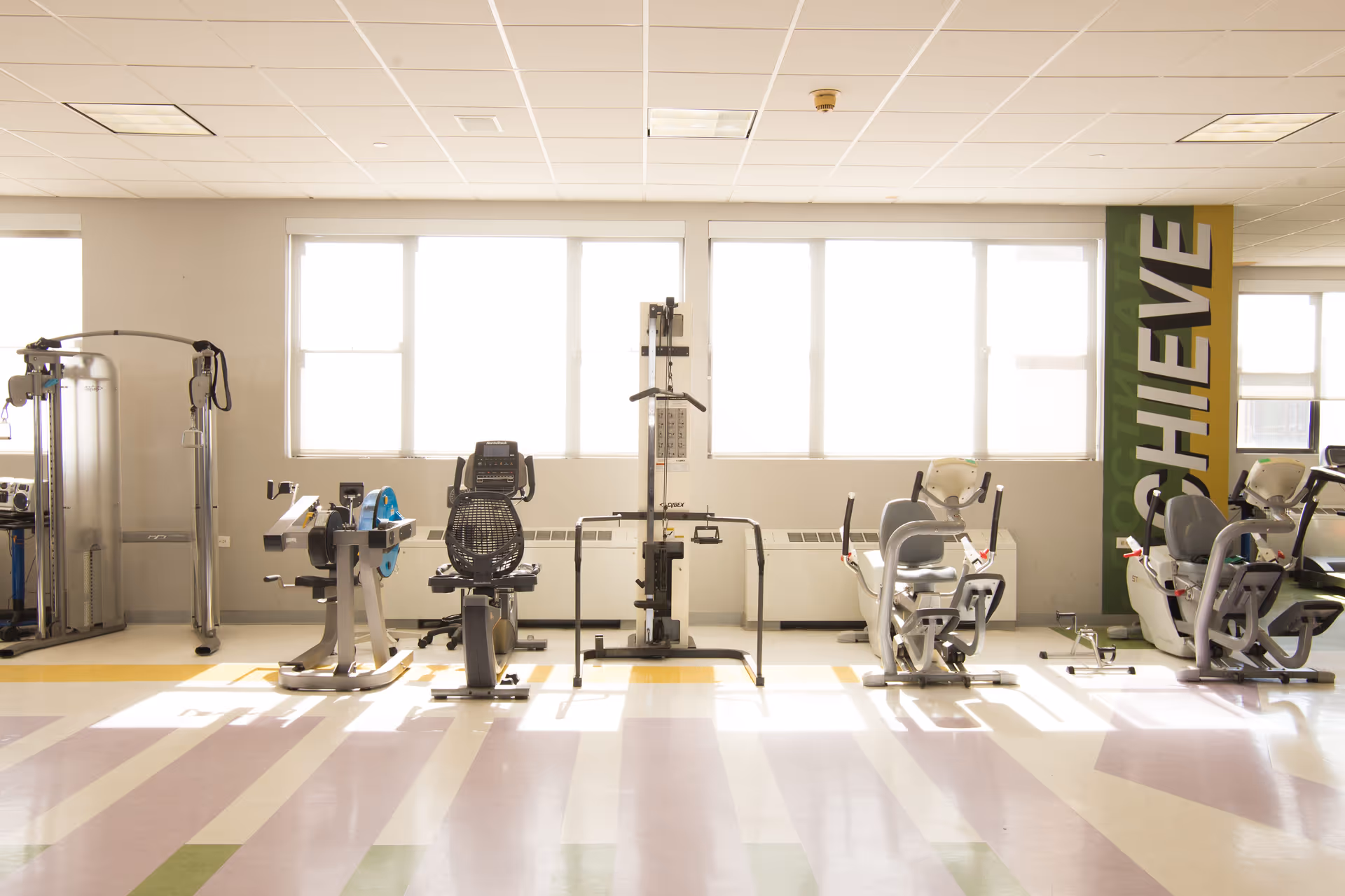 Sunlit fitness room with several stationary exercise machines lined up in front of large windows and a vertical 'ACHIEVE' sign.