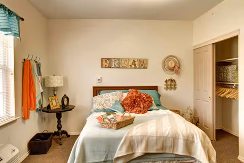Cozy decorated bedroom with a made bed, wall art reading 'DREAM', side table, window, and an open closet.