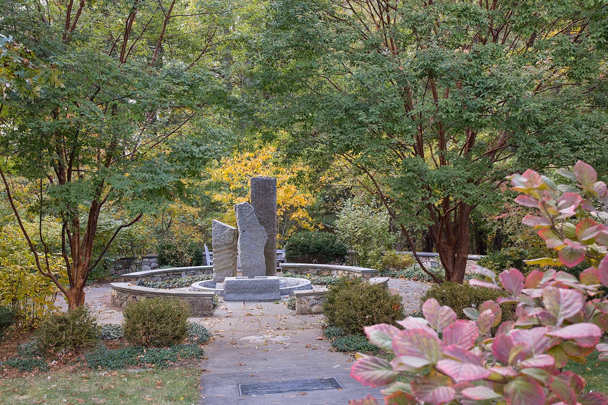 A peaceful outdoor garden area with a circular stone bench surrounding three vertical stone sculptures. The garden is lush with green trees and bushes, with some leaves showing autumn colors. A paved pathway leads to the stone sculptures.