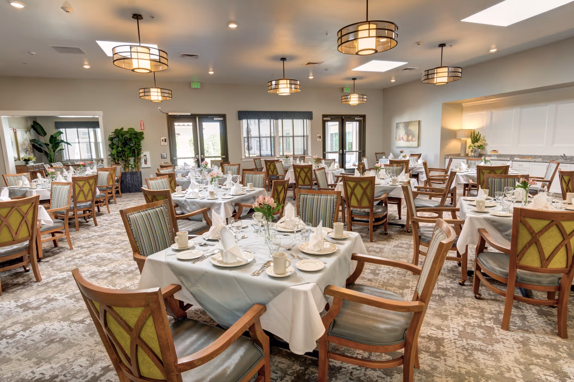 A spacious dining room with multiple tables set for a meal, each table covered with white tablecloths and neatly arranged with plates, cups, napkins, and cutlery. The room features wooden chairs with green and striped upholstery, modern ceiling lights, large windows, and doors letting in natural light. There are plants and decorative elements around the room, creating a welcoming atmosphere.