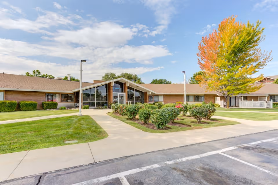 Single-story brick senior living building with a central glass entrance, landscaped lawn and a tree with autumn foliage.