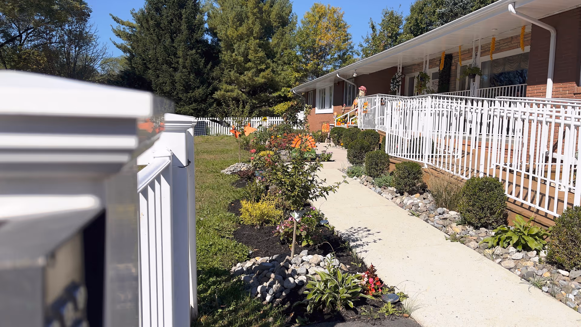 A paved walkway leading to the entrance of a single-story brick building with a white railing along the ramp. The walkway is bordered by landscaped garden beds with small bushes, plants, and flowers. Trees and a white picket fence are visible in the background under a clear blue sky.