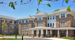 Front exterior of Perryman Station, a three-story senior living building with stone and siding facade, covered portico entrance, and landscaped grounds.