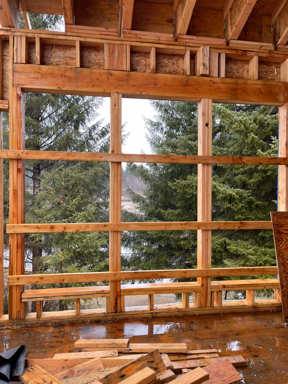 Interior view of a wooden framed wall under construction with large open window bays looking out to evergreen trees and scattered lumber on a wet floor.