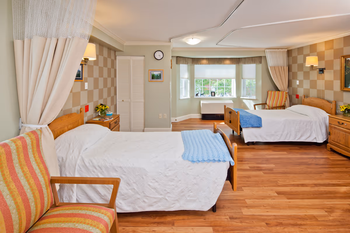 A bright and clean nursing facility room with two single beds covered in white bedspreads and blue blankets at the foot. Each bed has a wooden headboard and is accompanied by a wooden nightstand with a lamp and flowers. There are two striped armchairs, one near each bed. The room has wood flooring, a large window with a view of greenery, and light-colored walls with a checkered pattern behind the beds. A clock and framed pictures hang on the walls.