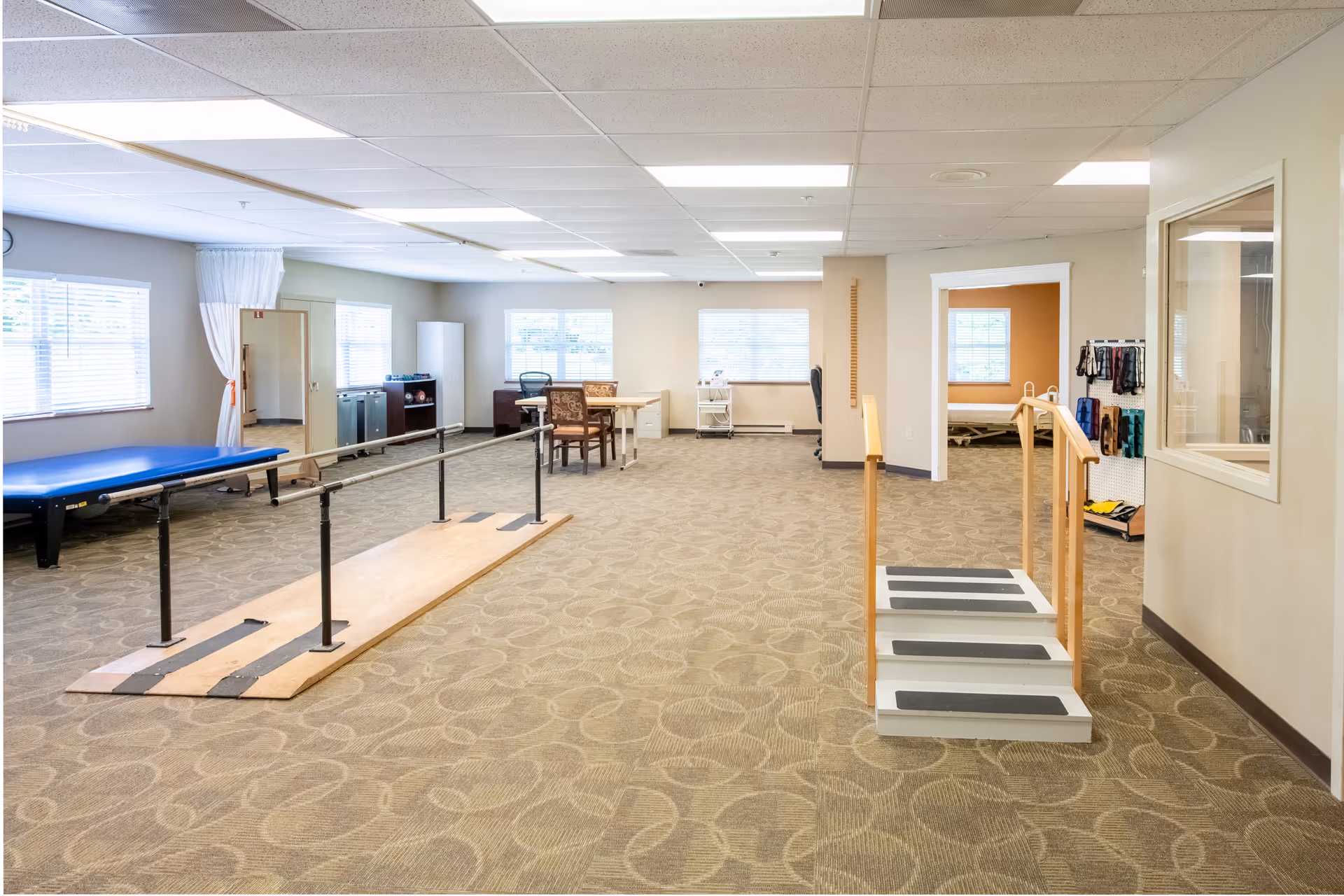 A spacious rehabilitation room with parallel bars for walking exercises, a small set of stairs with handrails, a blue therapy table, chairs, and windows letting in natural light. The room has beige walls and carpeted flooring with a patterned design.