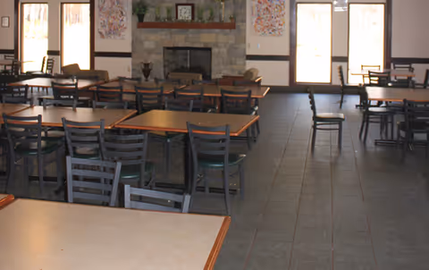 A spacious dining room with multiple tables and chairs arranged neatly. The room features large windows allowing natural light to enter, and a stone fireplace with decorative items on the mantel. The floor is tiled and the walls have framed artwork.