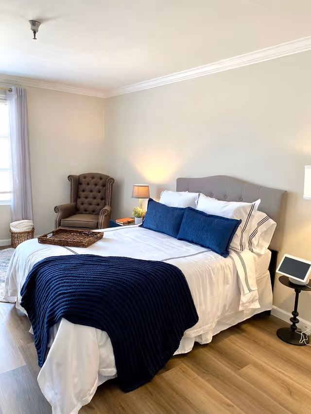 A neatly made bed with white sheets, two large blue pillows, and a dark blue knitted throw blanket at the foot. To the left of the bed is a brown tufted armchair near a window with light curtains. A small nightstand with a lamp and a small plant is next to the bed. The room has light-colored walls and wood flooring.